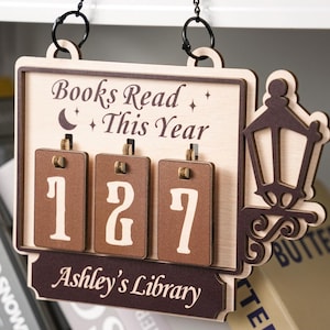 Könnte beinhalten: Holzschild mit der Aufschrift "Gelesene Bücher dieses Jahr" und der Zahl "127". Das Schild hat eine dekorative Straßenlaterne und den Text "Ashley's Library". Das Schild hängt an einer Kette.