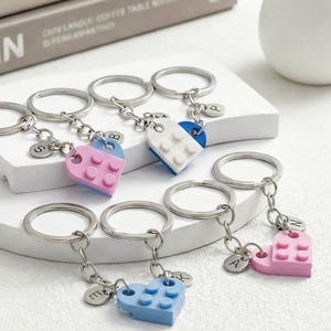 Puede incluir: Llaveros con dijes en forma de corazón hechos de bloques entrelazados en rosa, azul y blanco. Cada llavero tiene un anillo plateado y un pequeño dije redondo con una letra. Los llaveros se exhiben en una plataforma blanca.