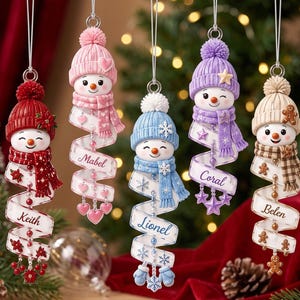 May include: Five hanging snowman ornaments with knit hats and scarves in red, pink, blue, purple, and beige. Each snowman features a spiraling white ribbon printed with the names Keith, Mabel, Lionel, Coral, and Belen, accented with beads and charms.