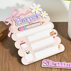 May include: A wooden first day of school sign with the name "Harper" in purple letters. The sign has spaces to write details about the school year, with a pink bow and daisy accents. A separate nameplate says "Sienna".