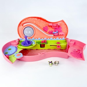 May include: A pink and orange toy playset with a garden theme, featuring a small pond, a purple spinning top, and two miniature animal figures. The playset is open, revealing interior details and decorative elements.