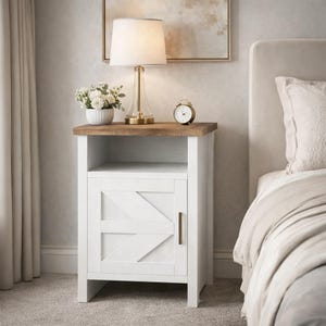 May include: A white nightstand with a wooden top and a single door featuring a geometric design. A gold-toned lamp, a small alarm clock, and a vase of white flowers sit on top. The nightstand is next to a bed with a cream-colored headboard.
