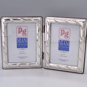 Può includere: Una cornice per foto argentata con cerniera e bordo intrecciato. Ogni sezione della cornice mostra un interno bianco con il logo "Pg Miani Venezia". La cornice è progettata per contenere due foto.