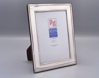 May include: A silver picture frame with a beveled edge and a dark wood backing. The frame has a label that reads "Pg Miani Venezia."