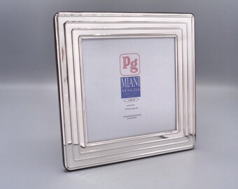 May include: A silver picture frame with a beveled edge and multiple layers. The frame has a small label with the text "Pg Miani Venezia" and a logo.
