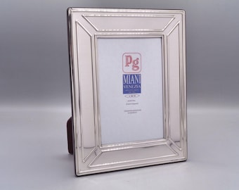 May include: Silver picture frame with a beveled edge and a raised border. The frame has a small label with the text "Pg" "Miani Venezia" and "Argento 925/1000".