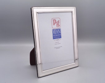 May include: A silver picture frame with a white mat and a silver border. The frame has a label that reads "Pg Miani Venezia Artigiano Veneto dal 1968".