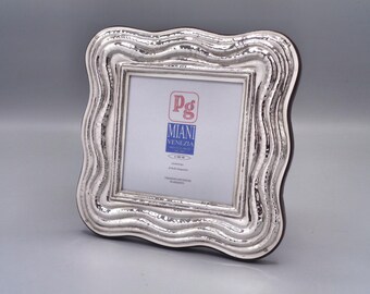 May include: A silver-colored picture frame with a wavy, textured border. The frame has a square opening and a dark brown backing. The frame has the text "pg Miani Venezia" on the front.