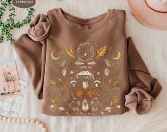 May include: A brown sweatshirt with a graphic print of mushrooms, crystals, birds, and leaves. The print is in shades of brown, yellow, and white.