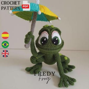 May include: A crocheted green frog with large, expressive eyes, holding a miniature umbrella with yellow, blue, and green panels. The text "FREDY Frog" is below the frog. The image also includes the text "CROCHET PATTERN".