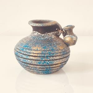 Peut inclure: Petit vase rond en céramique avec une surface texturée et une petite cruche attachée. Le vase présente des rayures horizontales bleues, dorées et noires. Le bord est brun foncé et la petite cruche est dorée.