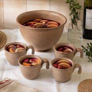 Peut inclure: Un grand bol à punch en céramique beige mouchetée et quatre tasses assorties, remplis d'un liquide rouge foncé contenant des tranches d'orange et de pomme. L'ensemble présente une finition rustique texturée et des anses incurvées.