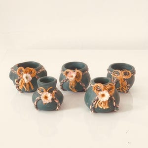 Peut inclure: Cinq pots en céramique turquoise ornés de raphia et de ficelle décoratifs. Chaque pot est orné d'une petite fleur blanche et est noué avec un nœud en raphia. Les pots sont disposés sur une surface blanche.