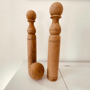 Op de afbeelding: Twee houten decoratieve objecten in de vorm van bowlingkegels en een houten bal. De objecten zijn lichtbruin met een afgeronde bovenkant en een cilindrisch lichaam. De bal is ook lichtbruin en bolvormig.