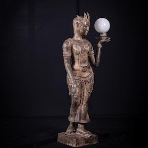 Op de afbeelding: Een bronzen beeld van een vrouw die een witte, bolvormige lamp vasthoudt. Het beeld is versierd met ingewikkelde details, waaronder een kroon en sierlijke kleding. De figuur staat op een rechthoekige basis tegen een zwarte achtergrond.