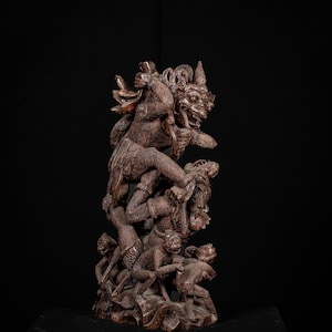 May include: A detailed, brown wooden sculpture depicting a mythological scene. The sculpture features multiple figures, including a large, fierce-looking creature atop smaller figures, all intricately carved with expressive details.