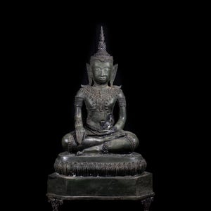 Può includere: Statua di Buddha seduta in bronzo con dettagli intricati. La figura è adornata con gioielli ornati e un'alta acconciatura. La statua è posizionata su una base decorativa su sfondo nero, mostrando la forma e l'artigianato della statua.