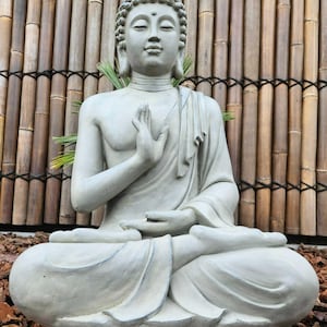 Può includere: Una serena statua di Buddha grigia in posizione di meditazione seduta. La statua presenta lineamenti dettagliati, una veste drappeggiata e mani in un gesto di pace. Lo sfondo include uno schermo di bambù e pacciame.