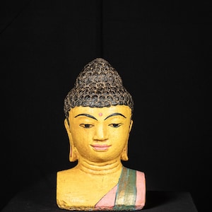 Puede incluir: Escultura de cabeza de Buda pintada a mano con una expresión serena. La cabeza está pintada de amarillo con cabello negro, rasgos delicados y un punto rosa en la frente. Una prenda drapeada en rosa, verde y dorado acentúa la escultura.