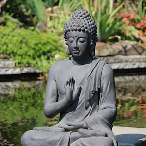 May include: A gray Buddha statue in a seated meditation pose, with hands in a gesture of prayer. The statue is outdoors, set against a backdrop of lush greenery and a tranquil pond. The sculpture has a serene expression.