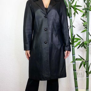 May include: A black knee-length coat featuring a classic collar, long sleeves, and a front button closure with three visible black buttons. The coat is constructed with vertical seam detailing along the front and sides, providing a structured fit.