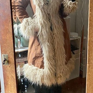 May include: A long brown coat featuring a prominent beige faux fur trim along the wide collar, the front opening, the sleeve cuffs, and the bottom hem. The coat has a smooth, solid-coloured brown exterior fabric and is worn over black trousers.