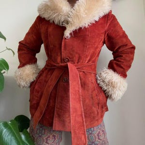 May include: A rust-colored suede jacket featuring a large cream-colored faux fur collar and matching faux fur cuffs. The jacket includes a front button closure with three visible buttons and a matching tie belt at the waist for a cinched fit.