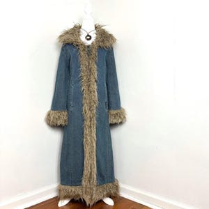 Könnte beinhalten: Ein langer blauer Jeansmantel mit einem dicken, zotteligen beigen Kunstfellbesatz am übergroßen Kragen, an der durchgehenden Vorderöffnung, an den Ärmelbündchen und am unteren Saum. Der Mantel hat eine strukturierte Silhouette.