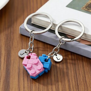May include: Two keychains with silver rings and chains. Each keychain has a small, round charm with a letter. The charms are attached to a heart-shaped design made of pink and blue blocks, resembling toy building blocks.