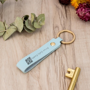 Könnte beinhalten: Hellblauer Leder-Schlüsselanhänger mit einem goldenen Schlüsselring. Der Schlüsselanhänger trägt den Text "Drive Safe Dad" und einen schwarz-weißen QR-Code.