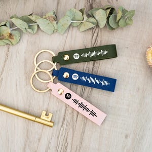 Könnte beinhalten: Drei Leder-Schlüsselanhänger mit goldenen Ringen. Jeder Schlüsselanhänger hat eine andere Farbe: grün, blau und rosa. Jeder Schlüsselanhänger hat ein Spotify-Logo und einen Barcode.