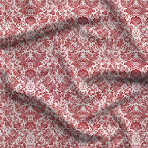 May include: A fabric featuring a repeating damask pattern in deep red on a white background. The material displays soft folds and creases across the surface, highlighting the intricate floral and scrollwork details of the printed design.