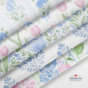 May include: A white fabric featuring a repeating floral pattern with pink tulips, blue bell-shaped flowers, and green foliage arranged in vertical stripes. The textile has a visible woven texture and includes a logo with the text AMERICAN FABRIC PRINTS.