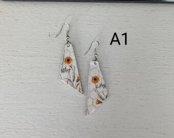 May include: A pair of earrings with a white background featuring a sunflower design. The earrings have a geometric shape with a honeycomb pattern and the text "Hello Sunshine". The earrings have silver-colored hooks.
