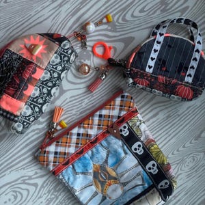 May include: Three Halloween-themed zippered pouches with various patchwork designs. The pouches feature skulls, pumpkins, and other spooky motifs. The pouches are made of fabric and have a black and white plaid pattern.