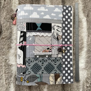 Può includere: Una copertina per diario in tessuto patchwork grigio e bianco con un motivo a teschi bianco e nero, un motivo a pois grigi e un motivo a nuvole grigie. La copertina ha un elastico rosa e un piccolo ciondolo dorato.