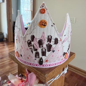 May include: A quilted Halloween crown with a spooky graveyard pattern, featuring ghosts, tombstones, and a jack-o'-lantern moon. The crown is adorned with orange buttons and a decorative border with the word "XOXOXO".