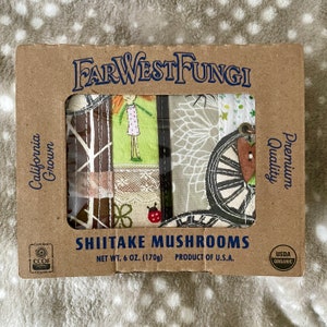 Puede incluir: Una caja de cartón marrón con una ventana que muestra un diseño de tela colorido. La caja contiene un paquete de champiñones Shiitake Far West Fungi. La caja está etiquetada con "California Grown", "Premium Quality", "Certified CCOF Organic" y "USDA Organic". El peso neto es de 170 g.