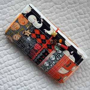 Könnte beinhalten: Ein Halloween-Notizbuch aus Stoff mit einem Patchwork-Umschlag mit Geistern, Kürbissen, Totenköpfen und Spinnweben. Das Notizbuch ist mit einem roten Gummiband gebunden und hat einen Quasten-Anhänger.