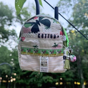 May include: A small quilted backpack pouch featuring a cream fabric with strawberry and butterfly prints. It includes a lace trim, a label reading COTTON, and a vertical tag labeled Home Made. A green tassel and strawberry charm hang from the zipper.