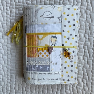 Könnte beinhalten: Ein handgefertigter Stoffbezug für ein Journal mit einem Patchwork-Design aus gelben, weißen und grauen Stoffquadraten. Der Bezug hat ein Bienen- und Sternenmotiv und den Text "Love you to the moon and back."