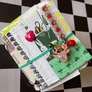 Puede incluir: Un pequeño libro hecho a mano con una cubierta blanca y parches de tela coloridos. Los parches presentan un fondo verde con un conejo blanco, una manzana roja, una abeja y un trébol verde. Un amuleto de reno rosa cuelga del libro.