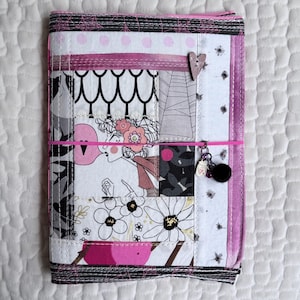May include: A rectangular fabric book cover featuring a patchwork design of white, black, and pink patterns, including floral motifs, a stylized character illustration, and spiderwebs. A thin pink elastic band secures the cover with a small charm.