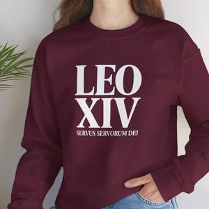 Può includere: Una felpa bordeaux con la scritta bianca "LEO XIV SERVUS SERVORUM DEI". La felpa è a girocollo e maniche lunghe. La persona indossa jeans blu.