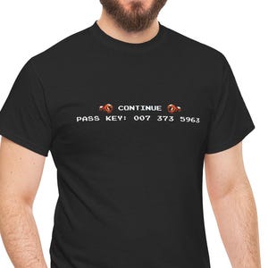 Op de afbeelding: Zwart t-shirt met de tekst "CONTINUE" en "PASS KEY: 007 373 5963" in een retro pixelstijl. Het shirt is gemaakt van zacht materiaal en is geschikt voor vrijetijdskleding.