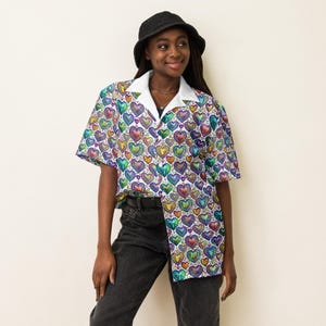 May include: A short-sleeved button-down shirt with a white collar and a vibrant pattern of colorful hearts. The shirt is paired with black jeans and a black bucket hat. The hearts are in shades of blue, green, purple, and yellow.