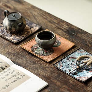 Könnte beinhalten: Drei quadratische Stoffuntersetzer mit komplexen Mustern, einer mit einer dunklen Keramik-Teekanne, einer mit einer kleinen dunklen Keramik-Teetasse und einer mit einer Metallschere mit umwickelten Griffen, platziert neben einem Buch mit chinesischem Text.