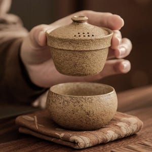 Op de afbeelding: Een beige keramische theeset met een deksel, bestaande uit een kleine, ronde kop en een bijpassende infuser met deksel. De infuser heeft kleine gaatjes voor theebladeren. De set staat op een bruine, bedrukte doek.