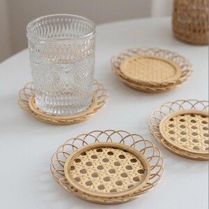 May include: A clear glass of water rests on a round, woven rattan coaster. Three more coasters are visible, displaying a natural, light brown hue and a decorative, scalloped edge. These coasters are designed to protect surfaces from drink condensation.
