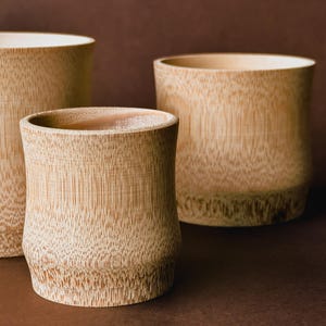 Può includere: Tre tazze cilindriche realizzate in legno di bambù color marrone chiaro, ciascuna caratterizzata da un distinto motivo a venature verticali e una parte superiore liscia e aperta. Le tazze variano leggermente in altezza e larghezza.
